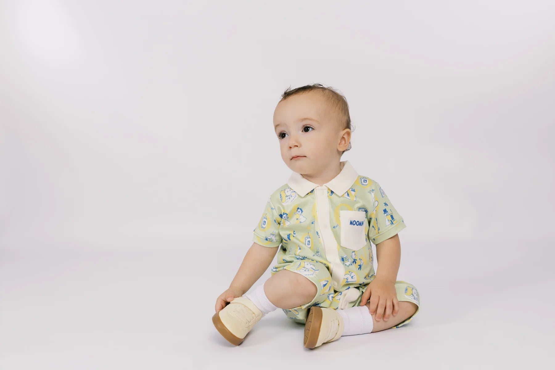 Baby in a green all-over Moomin print romper seated quietly beside the LOVE letter props.