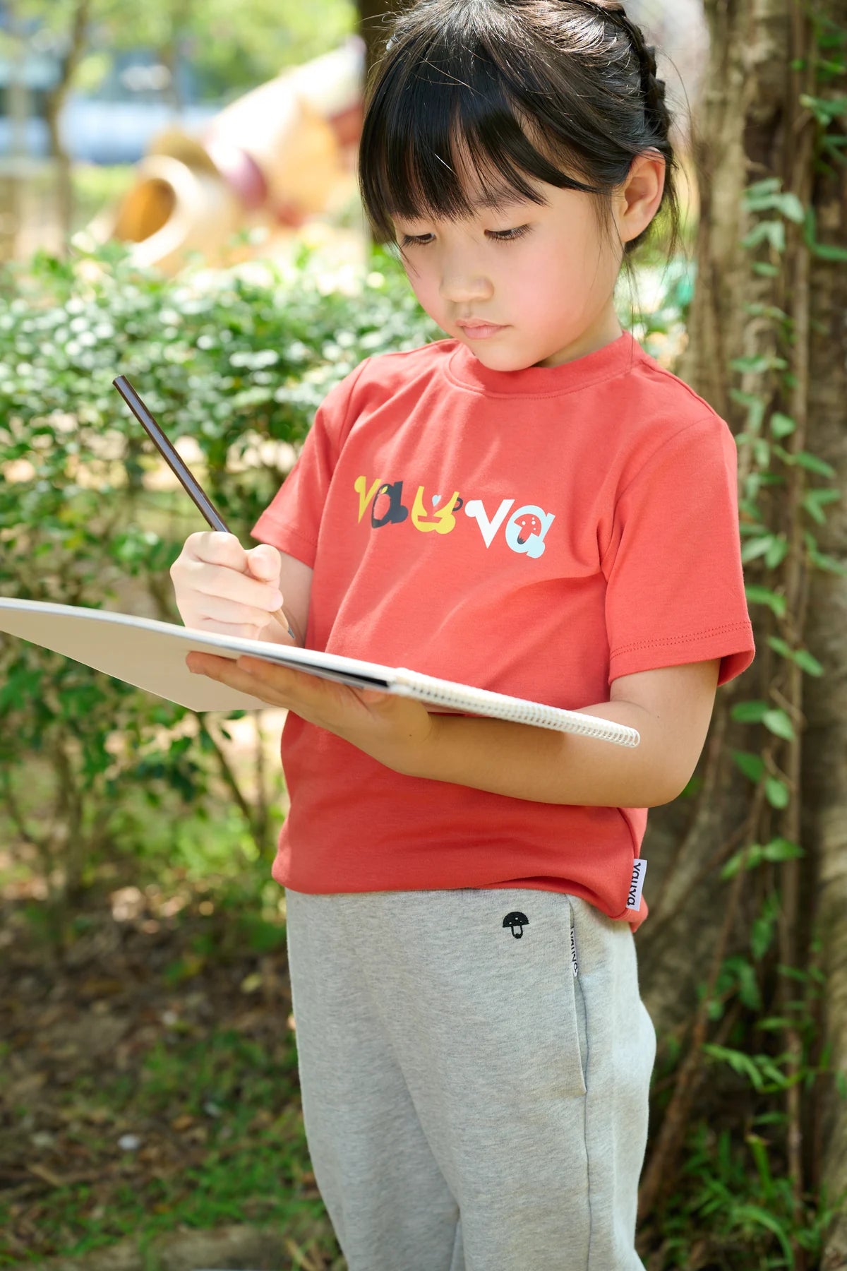 A child in grey everyday joggers and a red tee, sketching outdoors.