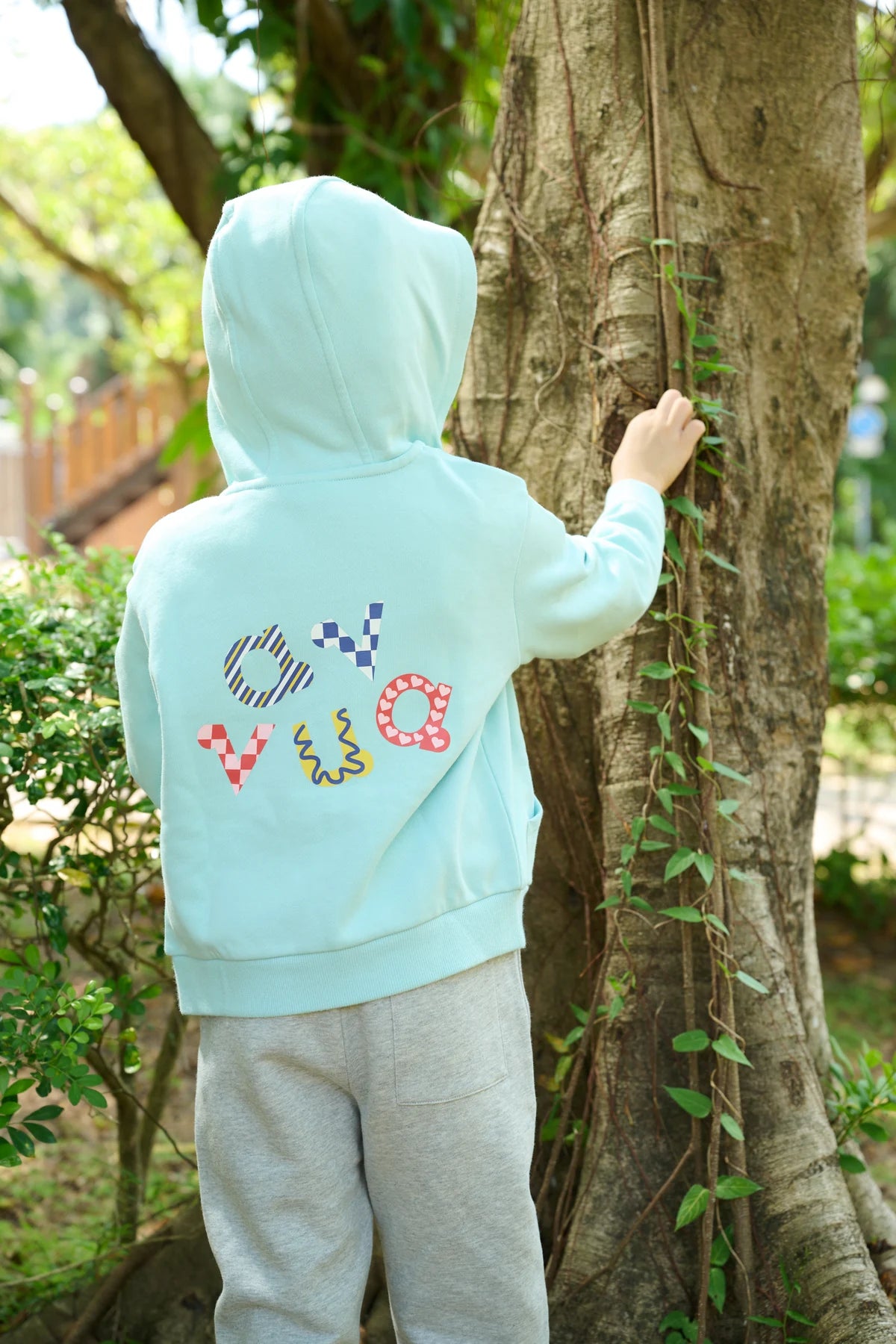 A pale teal hoodie with colourful embroidery, photographed from behind beside a tree.