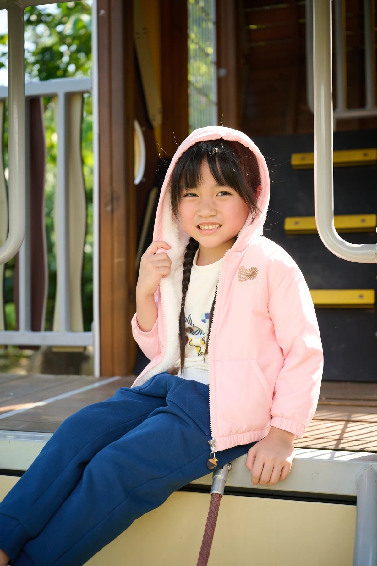 A child in a pink hooded jacket and navy joggers sitting in a playhouse doorway.