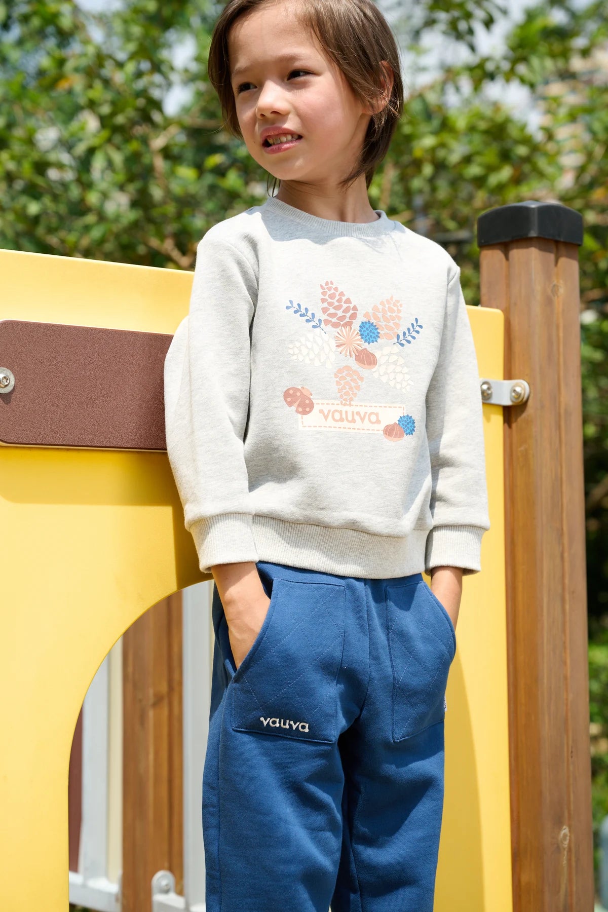 A child in a grey illustrated sweatshirt and navy joggers, standing by a yellow playhouse.