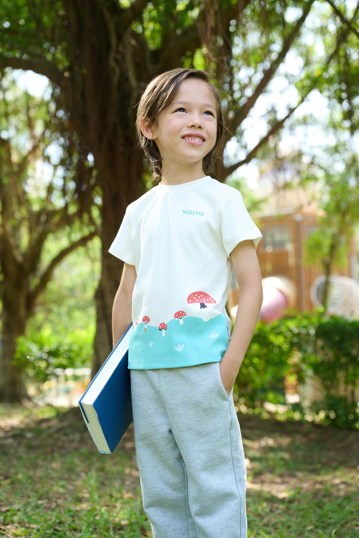 A child in a white and teal woodland-print tee, carrying a book outdoors.