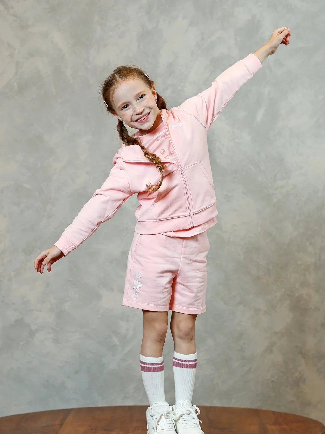 A child stretches her arms in the pink hoodie and coordinating embroidered shorts.