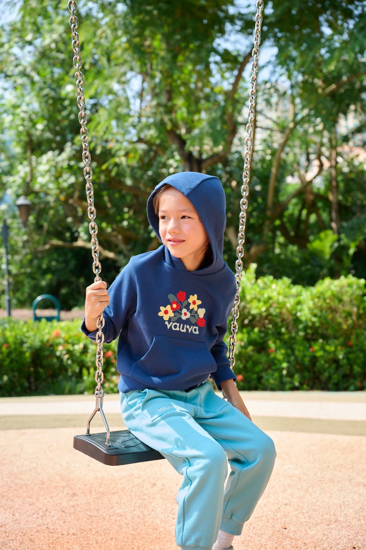 A child in a navy zip-up hoodie and teal joggers sitting on a swing.