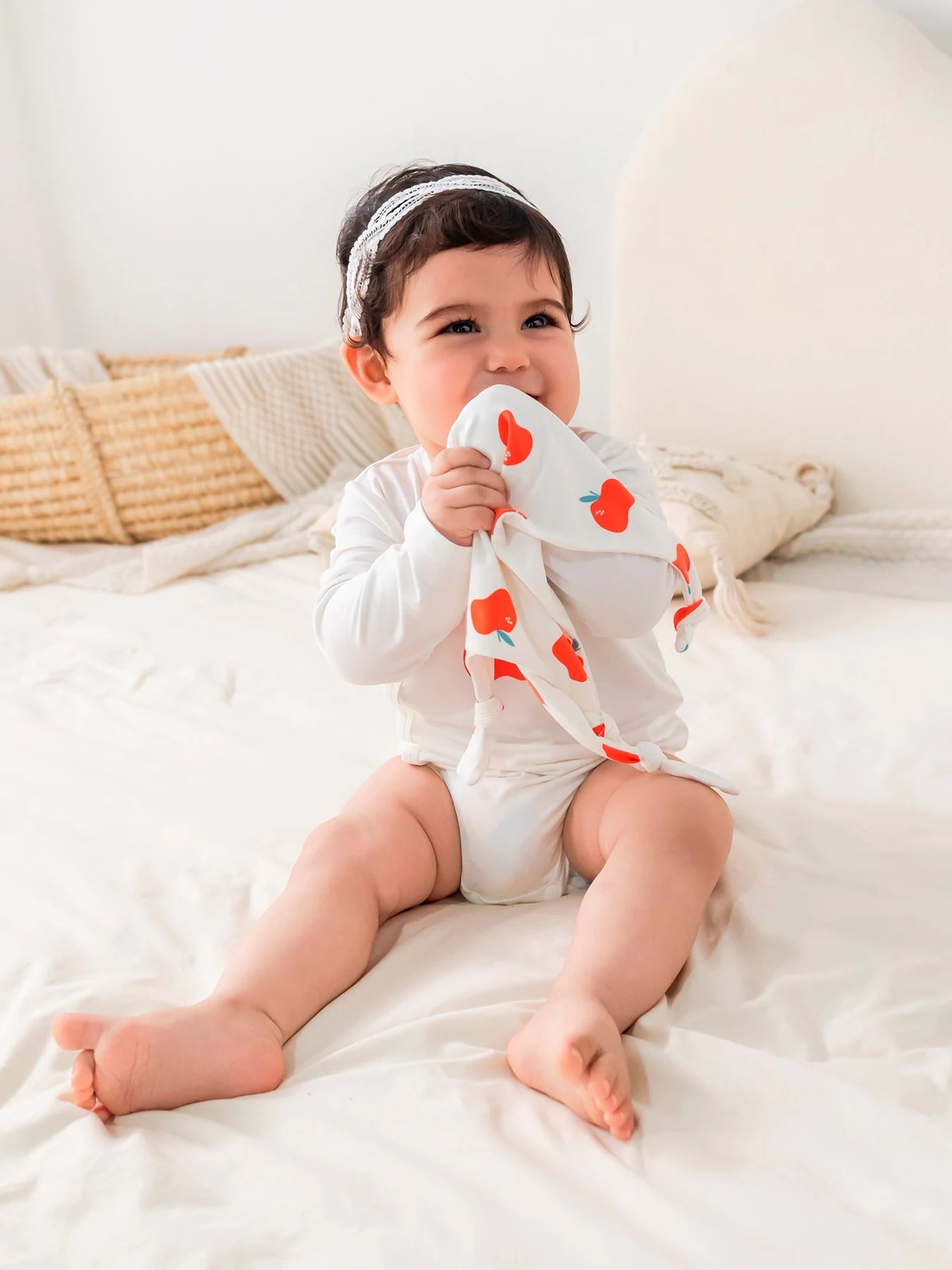 A baby seated on cream bedding in an apple-print long-sleeved bodysuit.