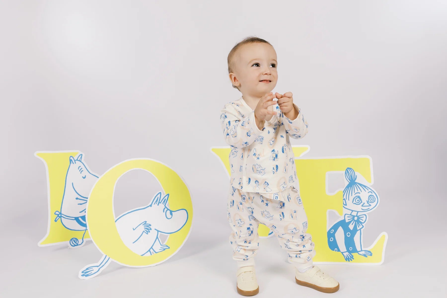 Baby standing in an ivory all-over Moomin print long-sleeved top and matching trousers.