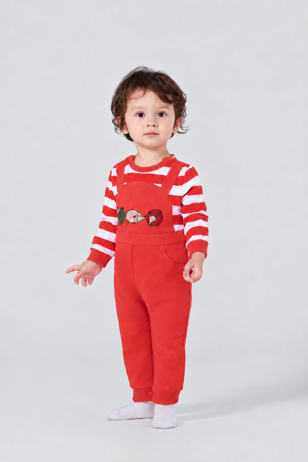 A child wearing red Vauva overalls with hedgehog appliqués and a striped shirt.