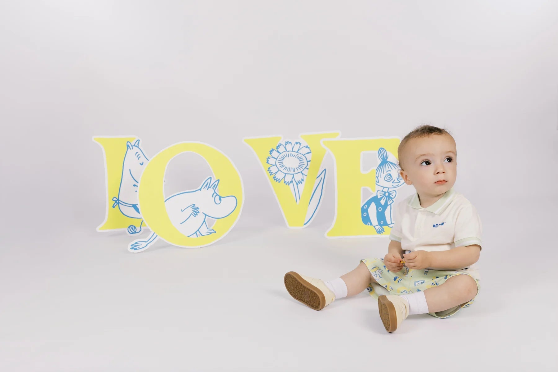 Baby in an ivory Moomin graphic T-shirt and green printed shorts, seated beside LOVE letter props.