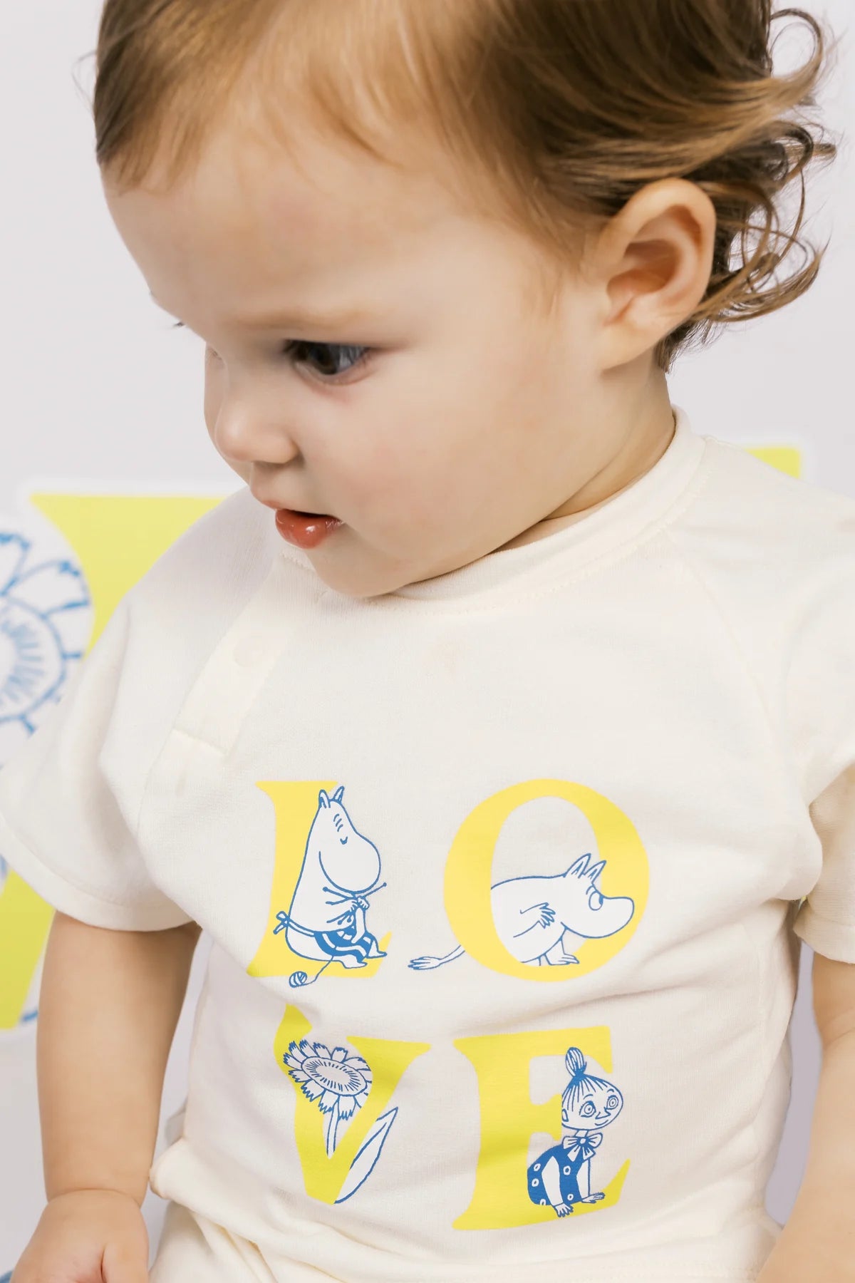 Close portrait of a baby looking down at the yellow LOVE letters and blue Moomin illustrations on an ivory top.