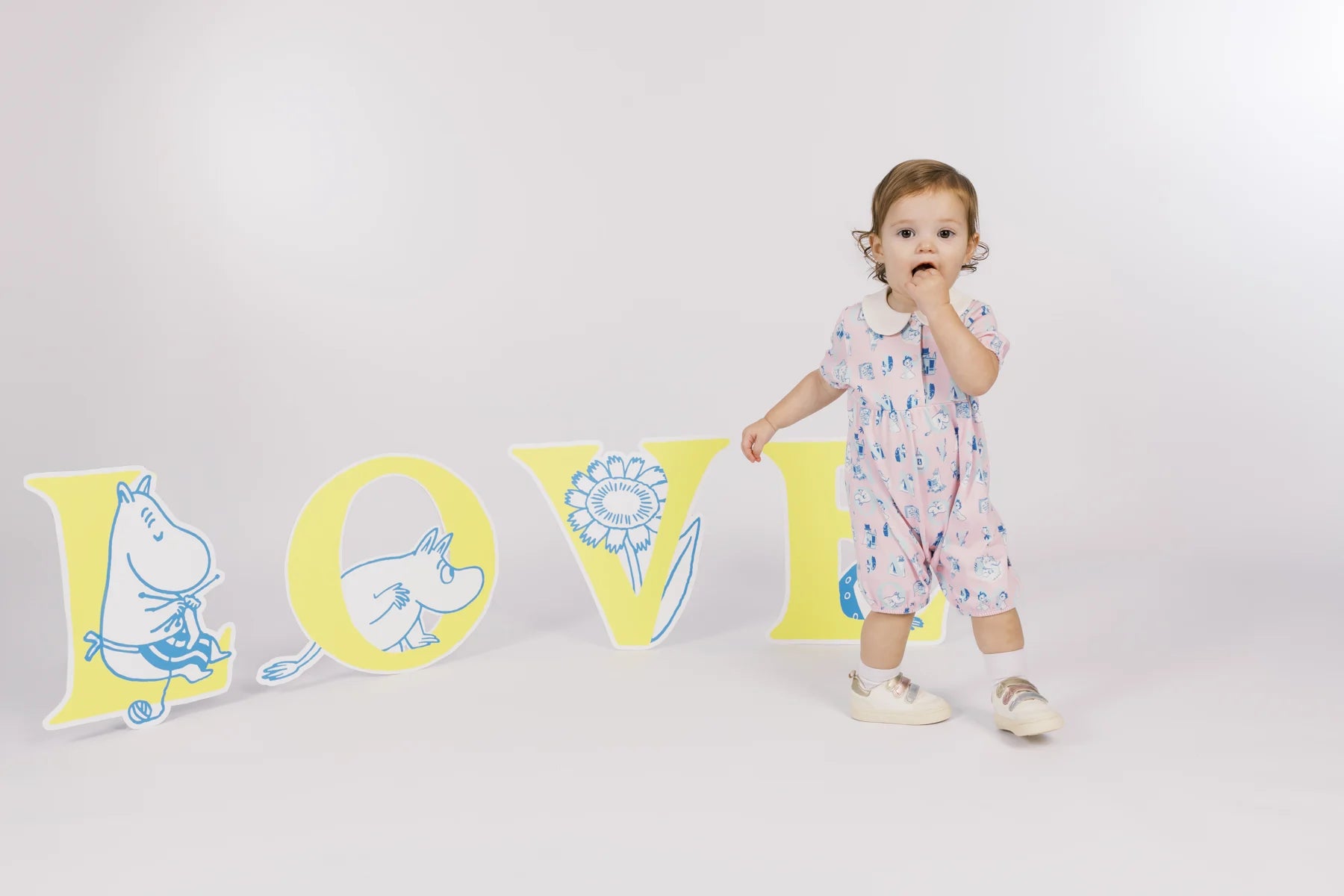 Baby in a pink all-over print Moomin romper beside oversized LOVE alphabet props.