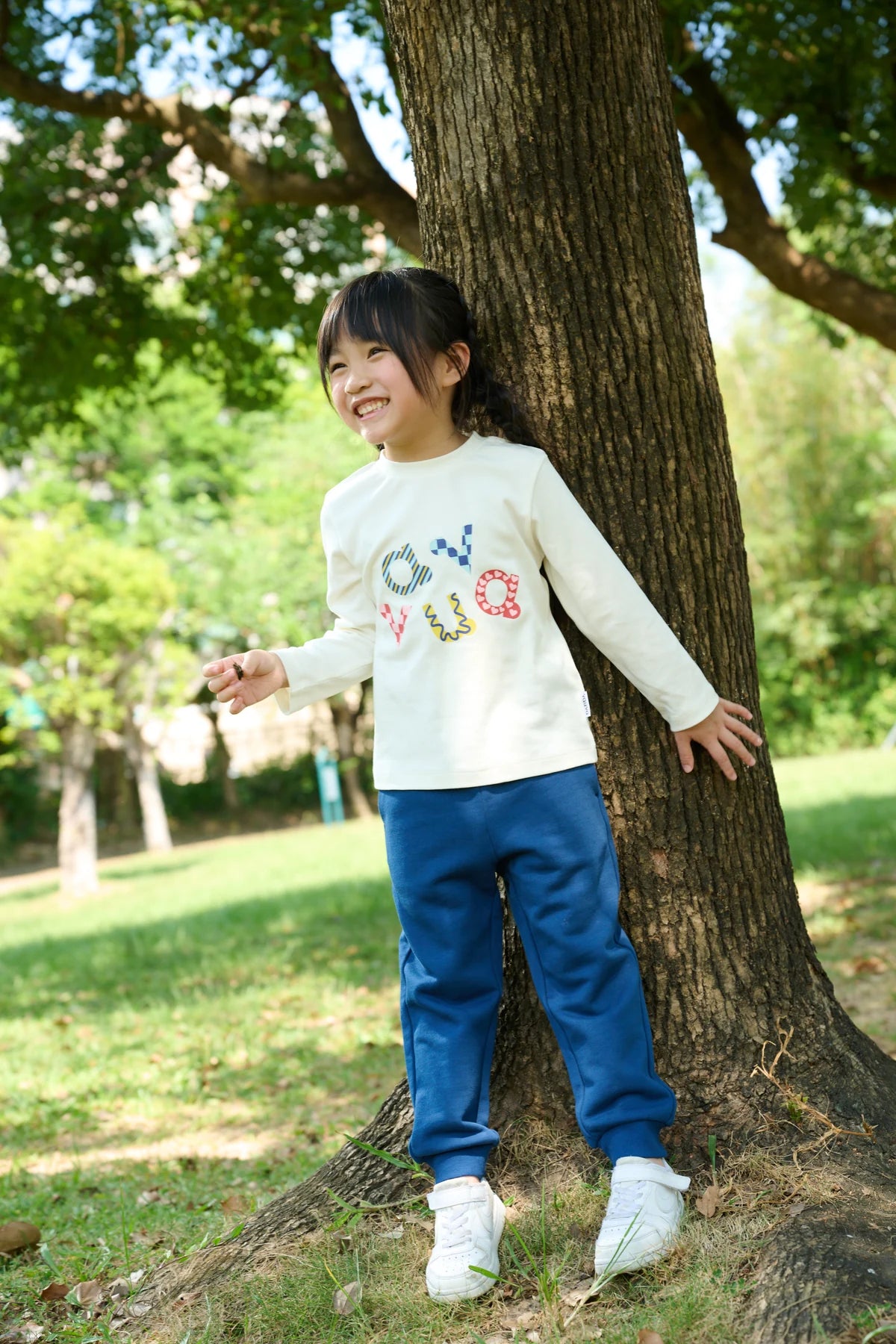 A child in a white colourful letter-print long-sleeve tee and blue joggers, playing beside a tree.