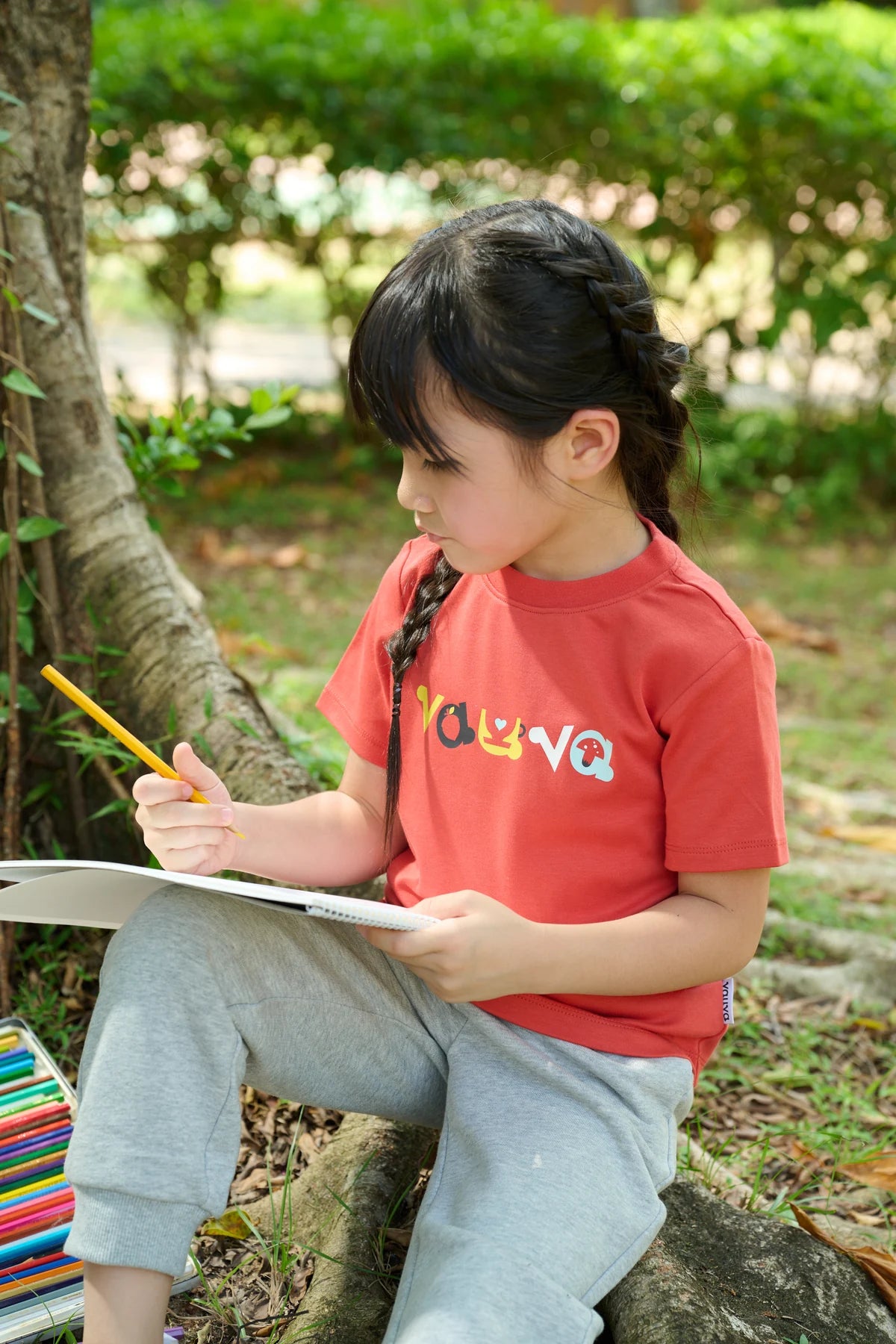 A child in a red Vauva print tee, drawing on a clipboard outdoors.