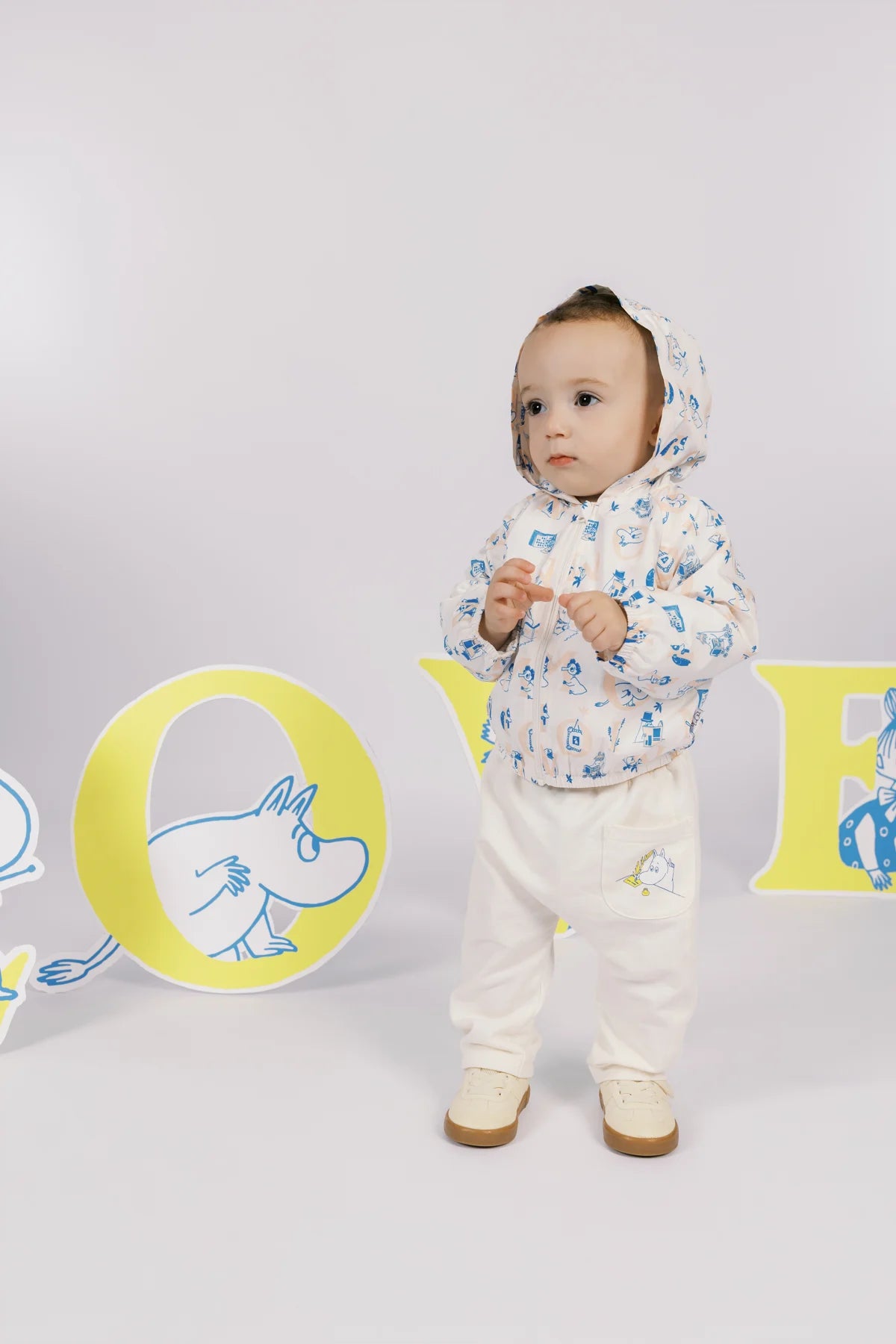 Baby wearing an ivory all-over Moomin print windbreaker and ivory trousers with a small print detail.