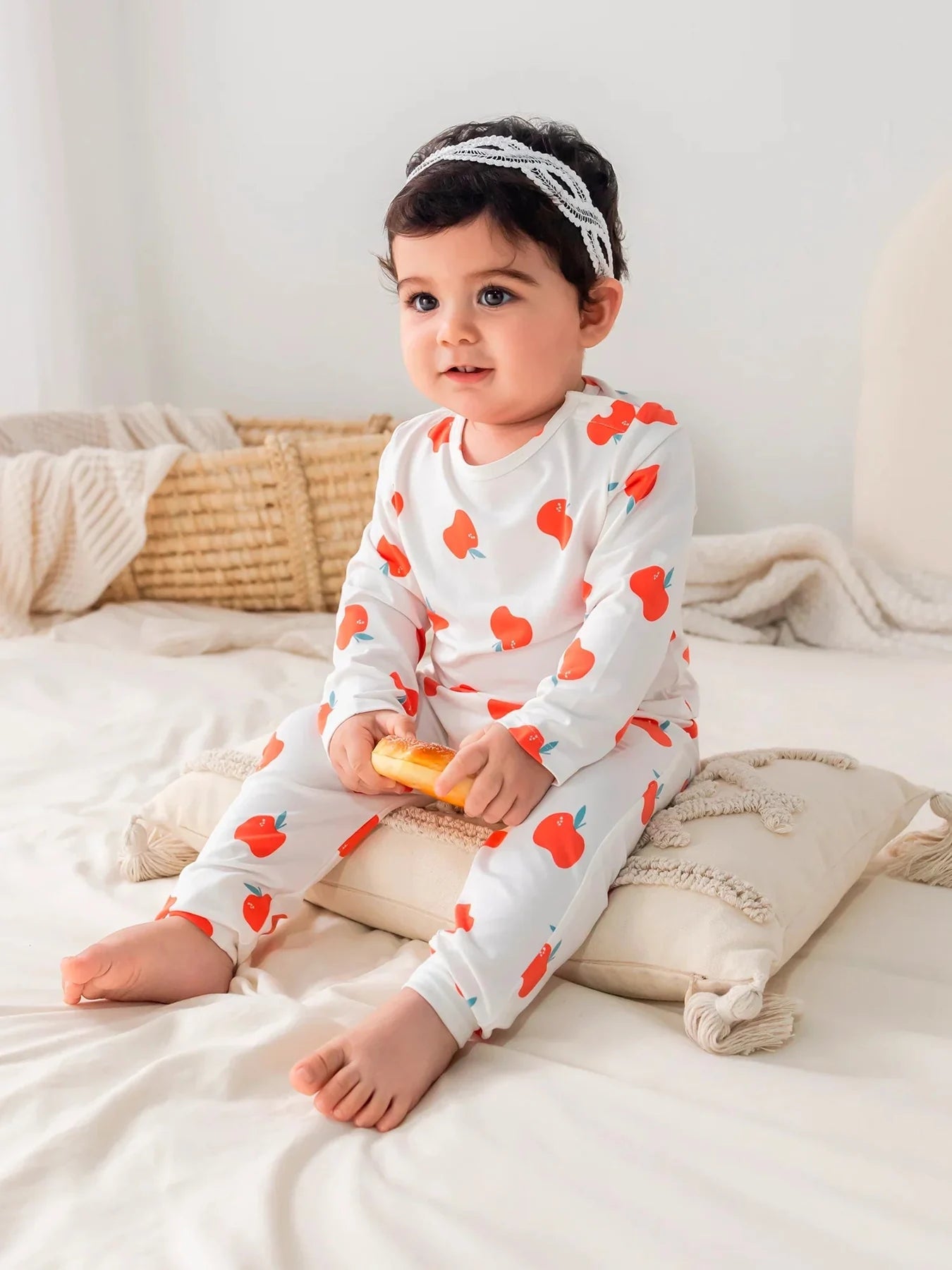 A baby seated on cream bedding in the apple-print top and coordinating bottoms.