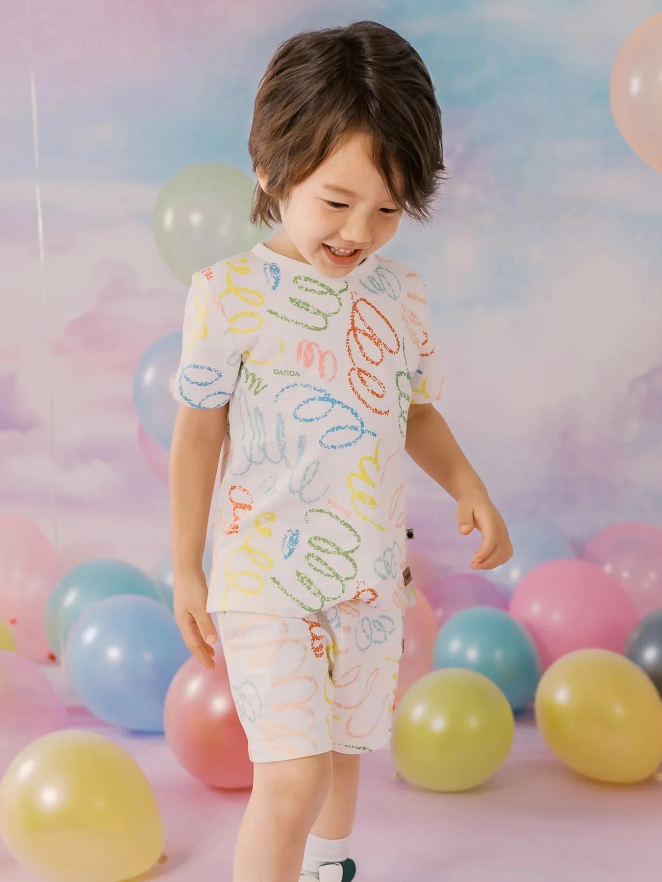 Child wearing a coordinated white scribble-print tee and shorts among pastel balloons.