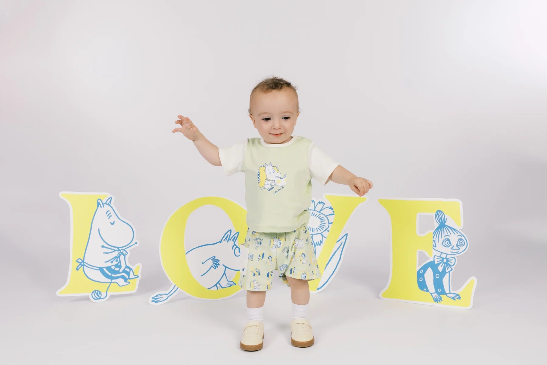 Baby wearing a seafoam green Moomin graphic T-shirt and green all-over print shorts, arms open.