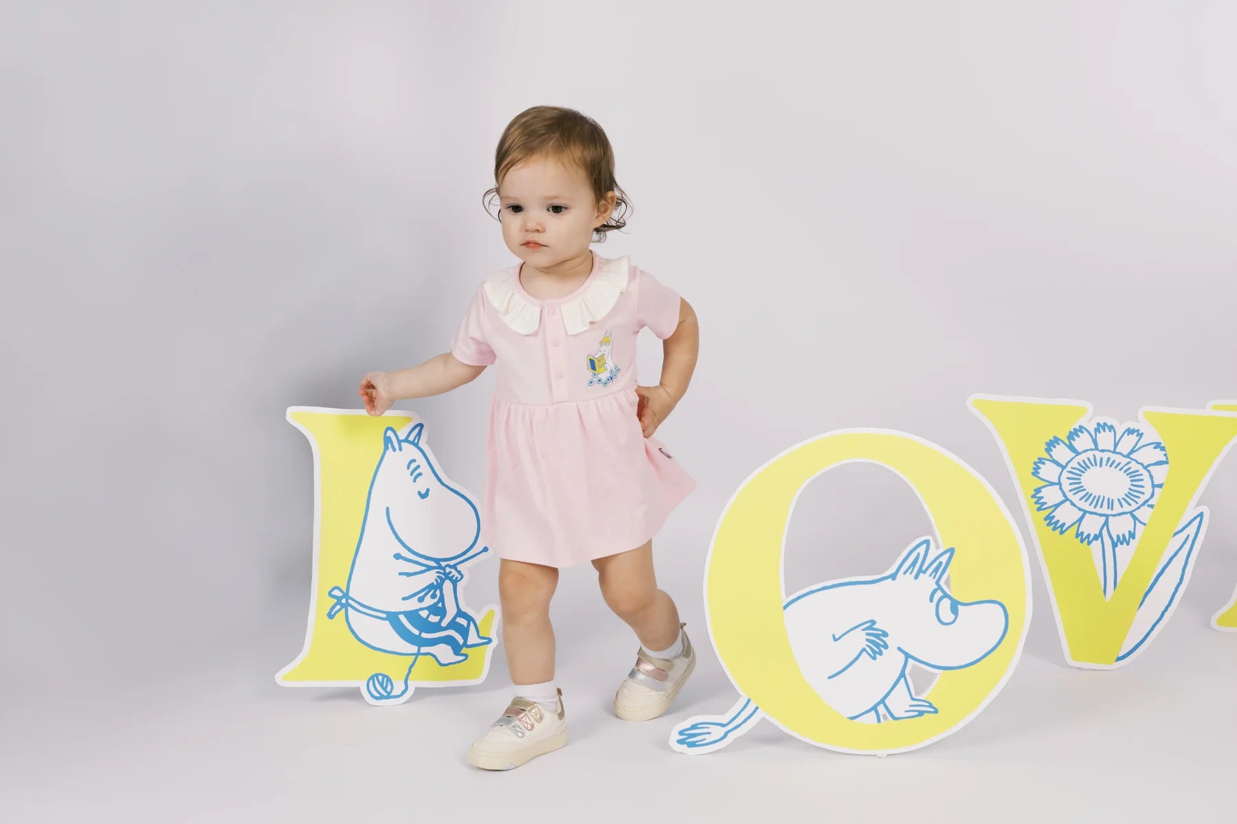 Baby in a blush pink Moomin bodysuit with a skirt-shaped silhouette, standing beside oversized LOVE letters.