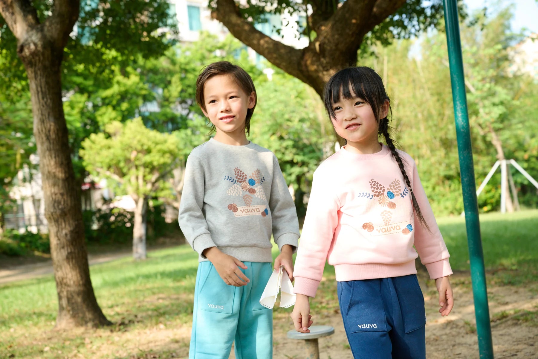 Two children standing together in a park, wearing grey and pink Vauva FW26 sweatshirts.